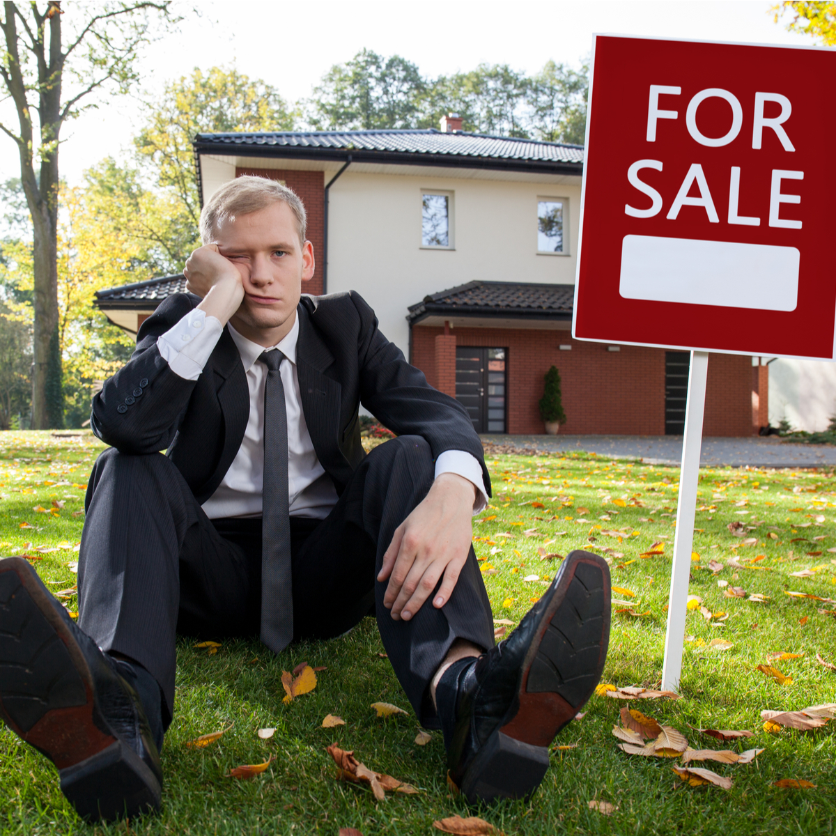 a realtor sitting on the grass next to a for sale sign in front of a house for sale